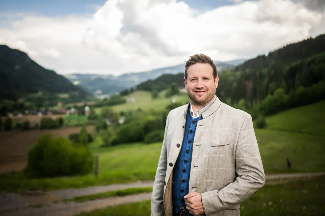 A man stands in a field, smiling with mountains and a cloudy sky in the background. He wears a light-colored jacket with a blue inner shirt and a vest.