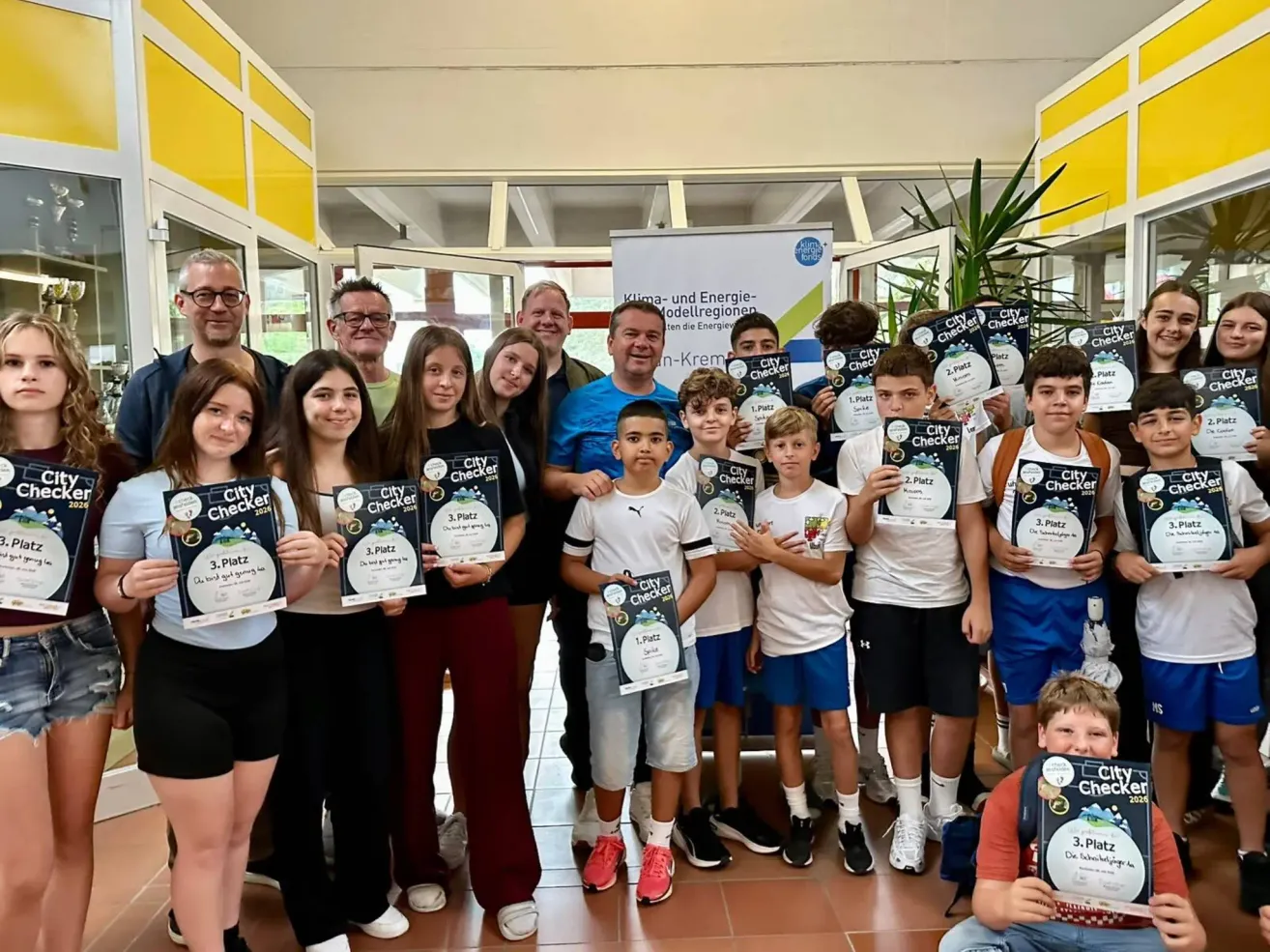 A group of people, including children, adults, and a few wearing glasses, are posing for a photo in a hall with certificates. They are holding certificates with the text 'City Checker' and '3. Platz'. Behind them is a banner with the same text.