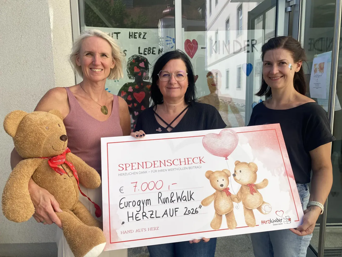 Drei Frauen präsentieren einen Scheck mit Teddybären. Sie stehen vor einer Glastür mit einem Plakat. Der Scheck ist für 7.000 Euro für den Eurogym Run&Walk 2026.