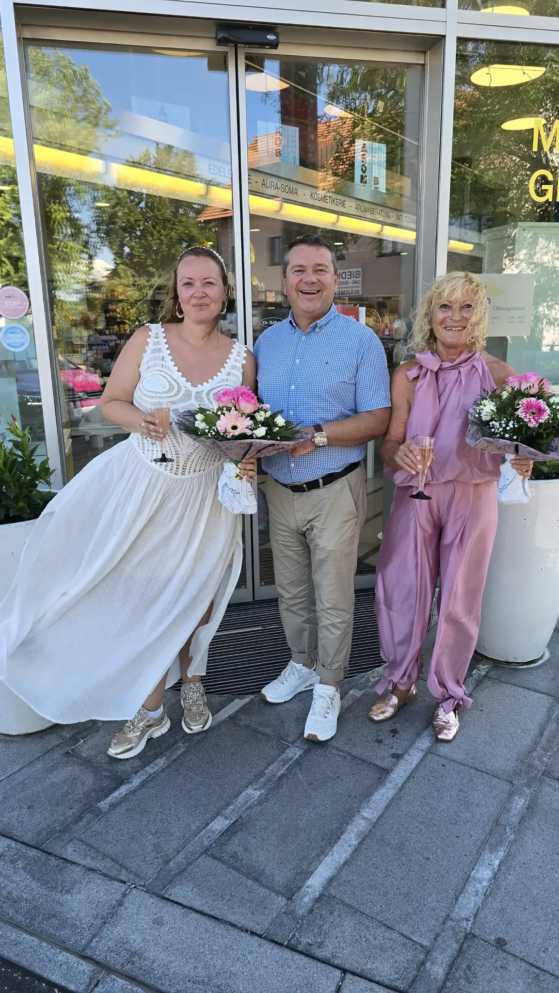Three people, two women and a man, stand outside a shop holding flowers. The women wear white and pink dresses. The man wears a blue shirt and khaki pants.