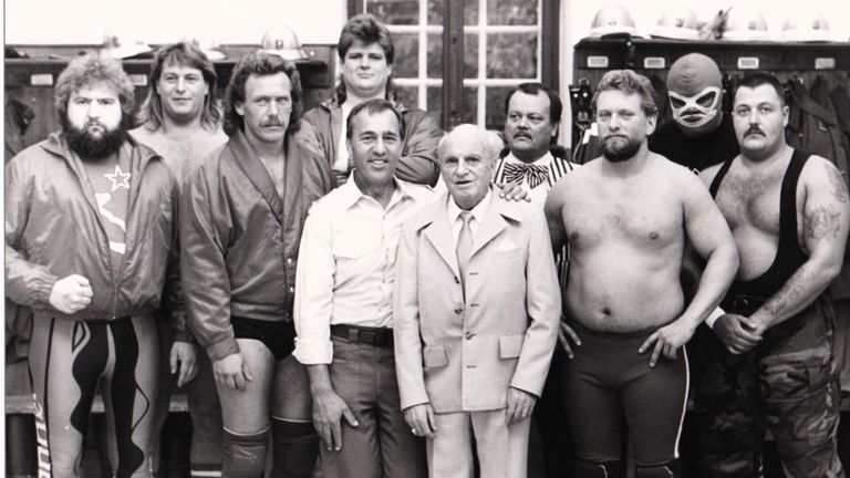 Ein Schwarz-Weiß-Foto zeigt eine Gruppe von Männern, die zusammenstehen. Der Mann in der Mitte trägt einen Anzug und eine Krawatte. Die anderen sind unterschiedlich bekleidet.