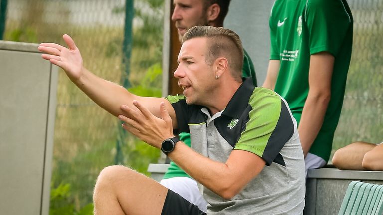 A man in green and gray sportswear is gesturing with his hands, possibly coaching a sports team. He is wearing a black wristwatch.