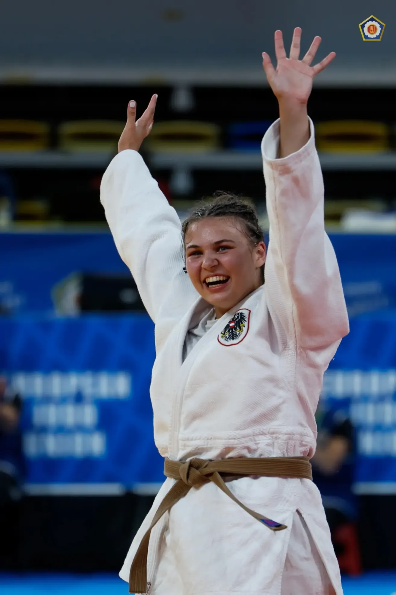 A woman in a white karate uniform and brown belt celebrates a victory. She raises both arms and smiles. A blue backdrop with white text is behind her.
