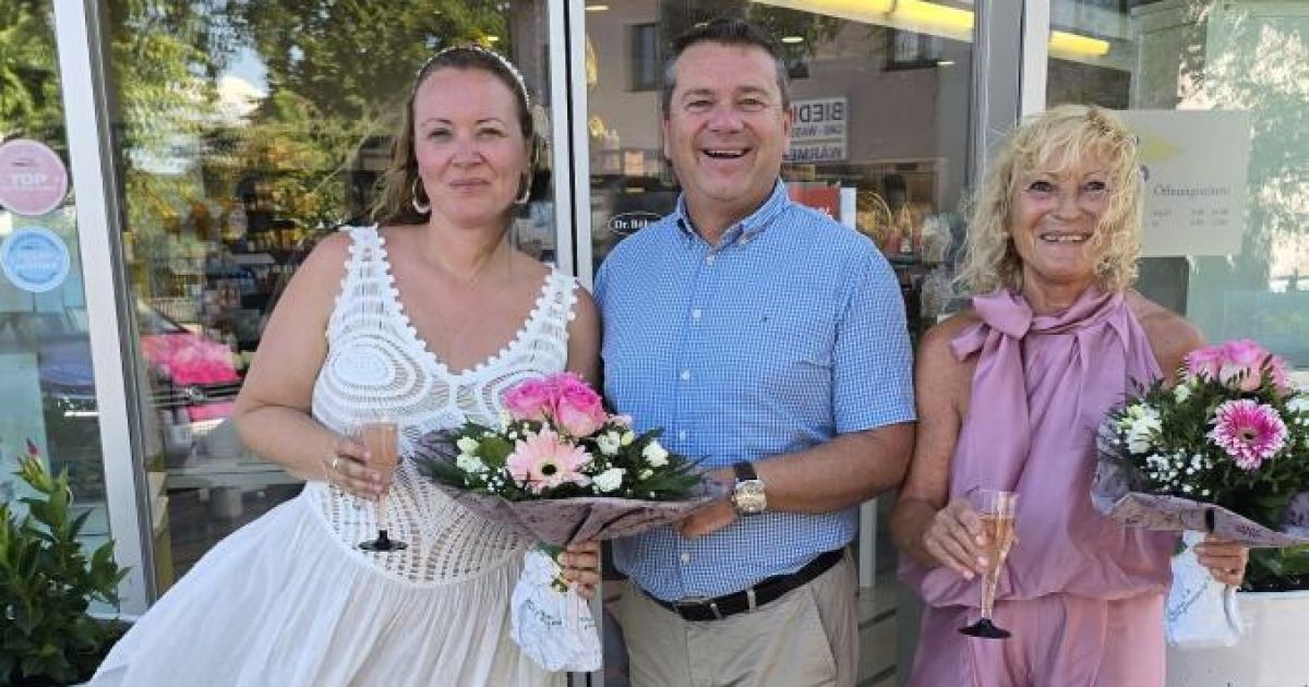 A woman in a white dress and two men in casual attire hold flowers and wine glasses, smiling outside a store.