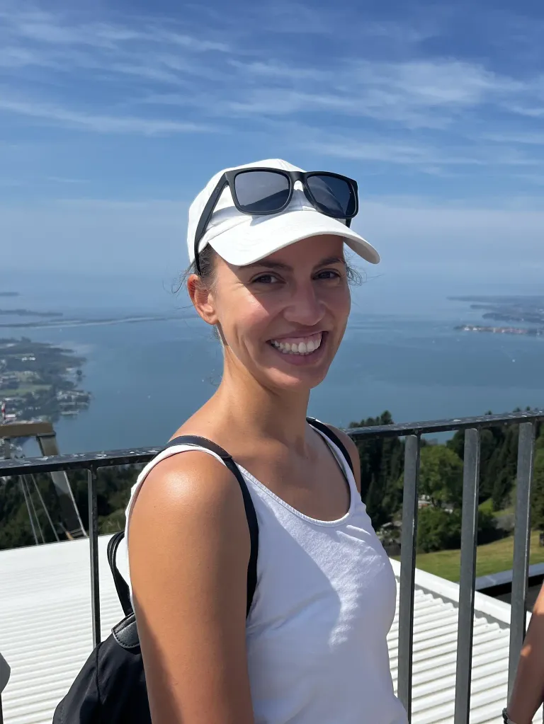 A woman smiles while wearing a white cap and sunglasses. She is standing on a balcony with a railing. Behind her is a view of the ocean and a city.