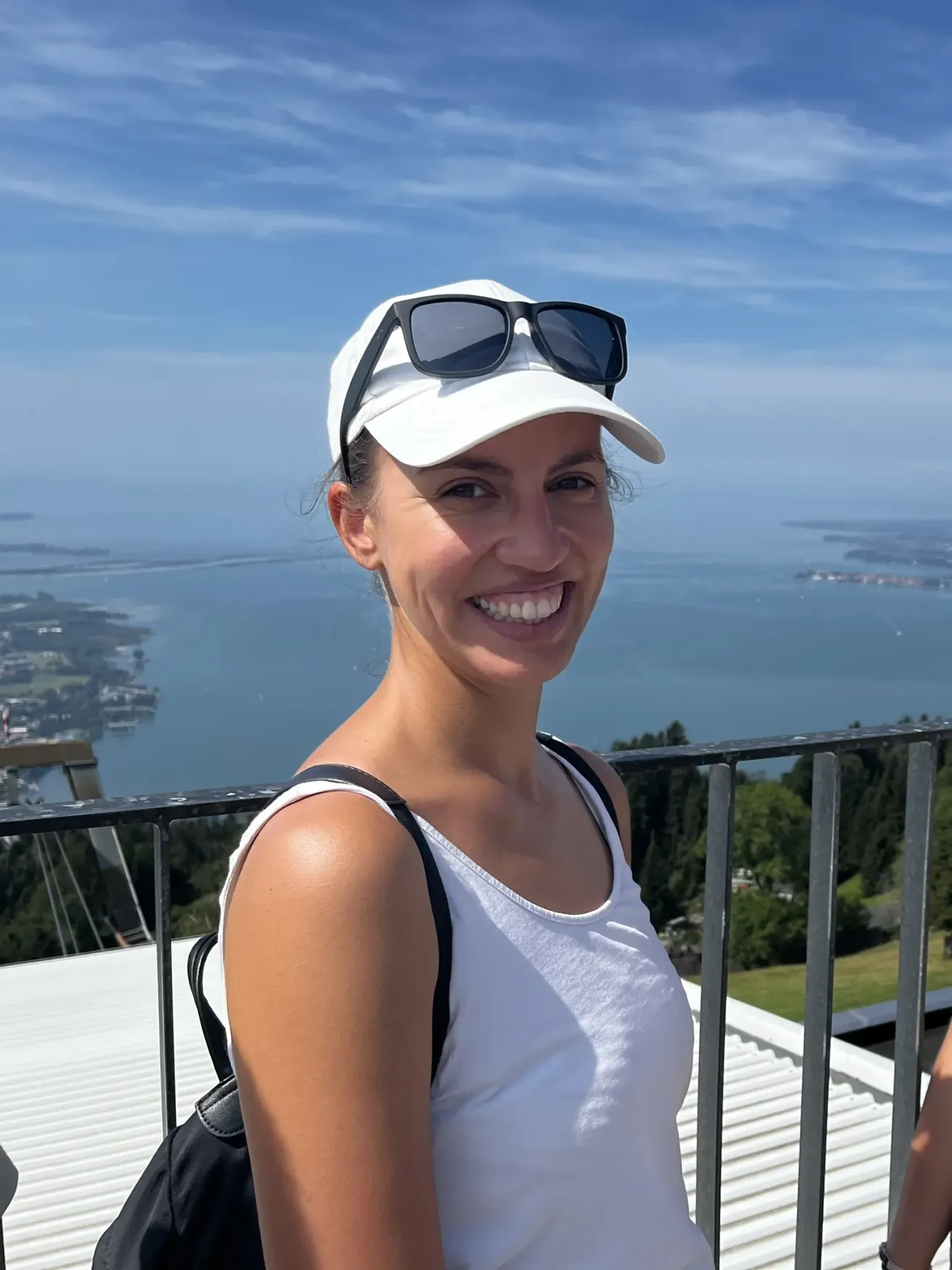 A woman smiles while wearing a white cap and sunglasses. She is standing on a balcony with a railing. Behind her is a view of the ocean and a city.