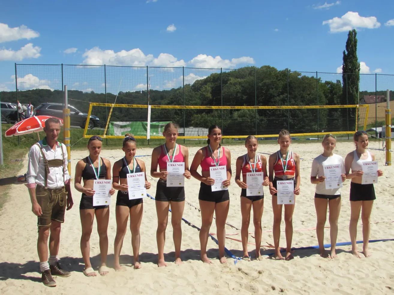 Seven women in swimwear stand on a beach holding certificates. A volleyball net and trees are in the background. A man in traditional clothing stands at the left.