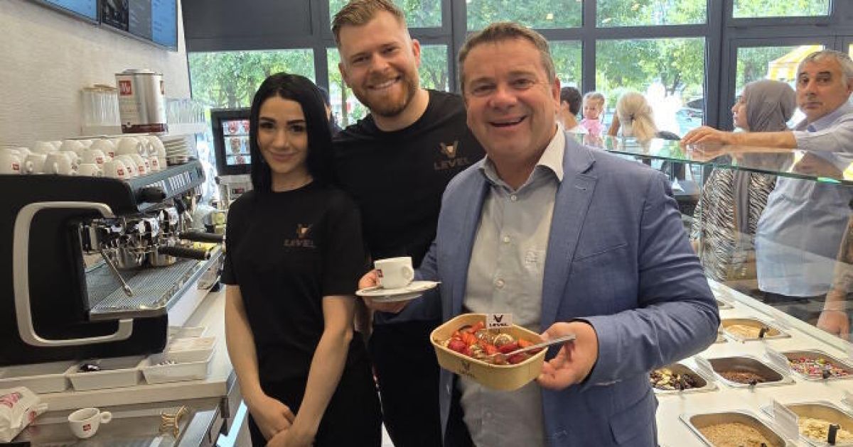 Three people pose for a photo in a coffee shop. A man in a suit holds food, while a woman and man in black shirts stand beside him.