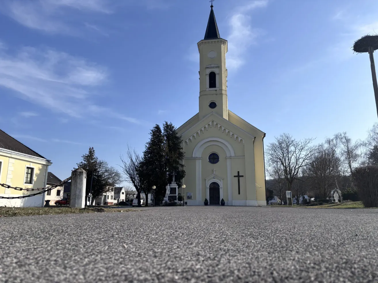 A yellow church with a steeple and a cross on its facade, surrounded by trees and a gravel road under a partly cloudy sky.