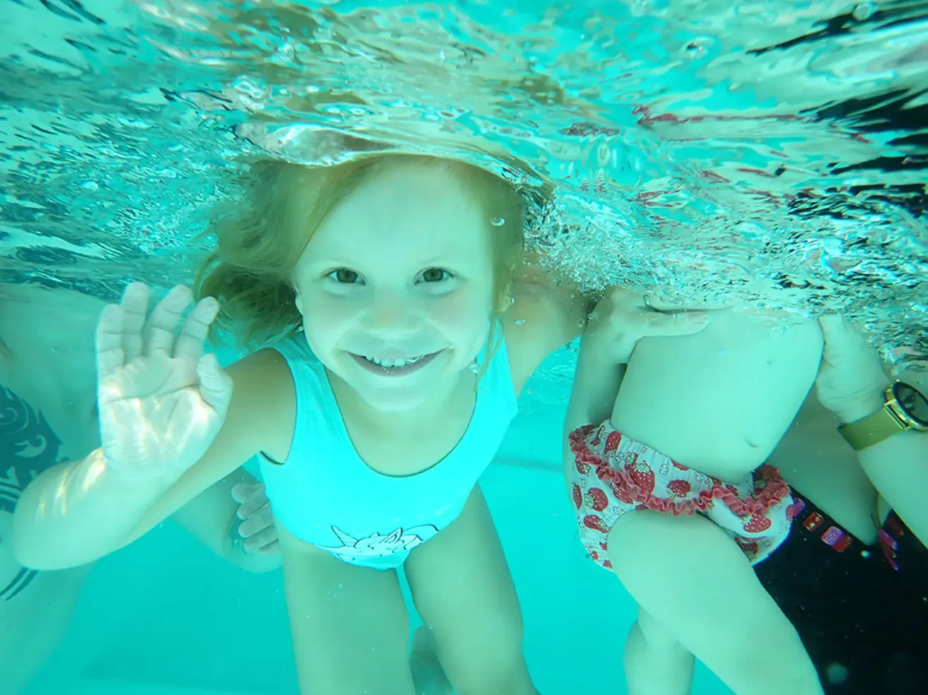 A young girl wearing a blue swimsuit is swimming underwater and smiling at the camera. She is holding a hand of another child.