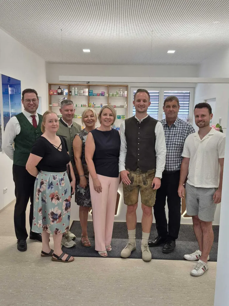 A group of people in a room, smiling and posing for a photo. A woman in a floral skirt stands on the left, and a man in shorts is on the right. Behind them is a cabinet with various items.