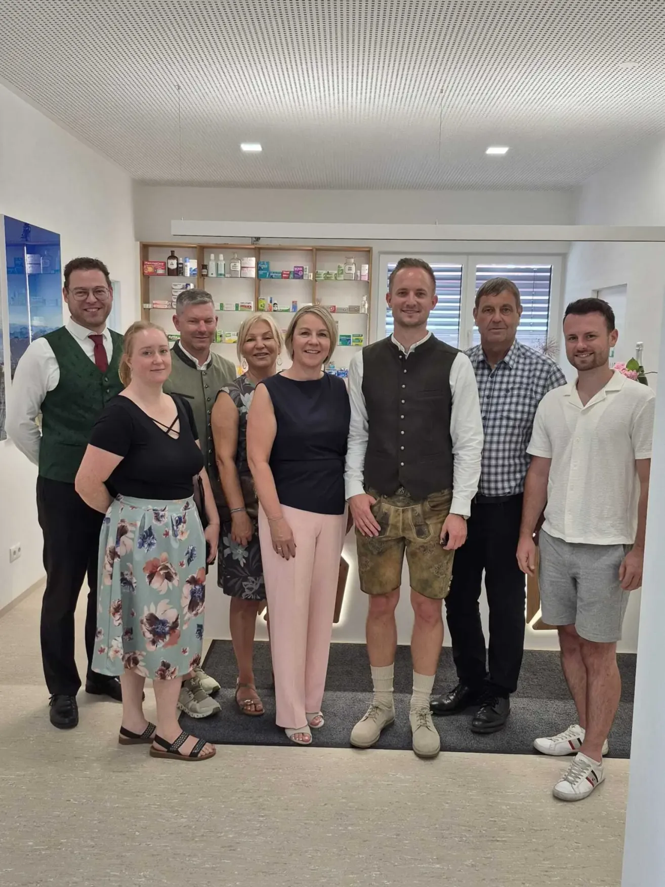 A group of people in a room, smiling and posing for a photo. A woman in a floral skirt stands on the left, and a man in shorts is on the right. Behind them is a cabinet with various items.