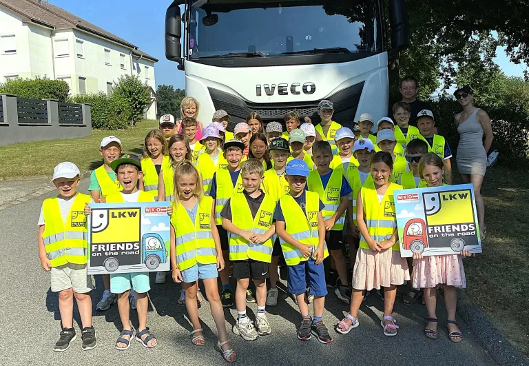 A group of children wearing yellow vests pose for a photo in front of an Iveco truck. They hold signs with the words 'LKKW' and 'FRIENDS on the road'.