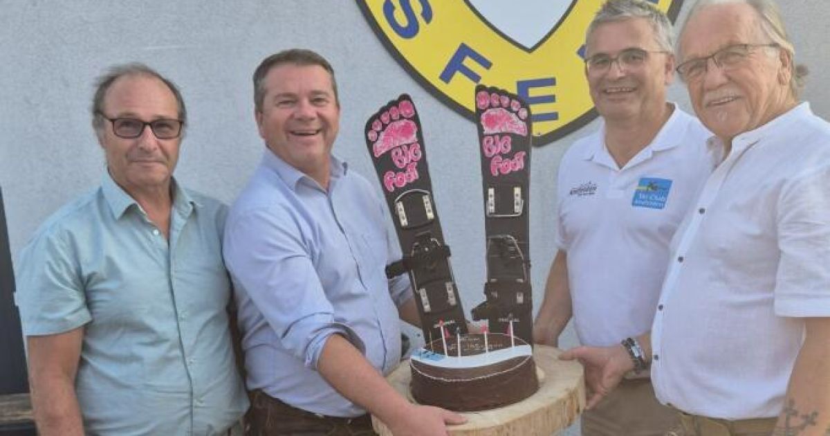 Four men stand around a chocolate cake with 'Big Foot' written on it, smiling and posing for a picture. The cake has two pink footprints and sits on a wooden surface.