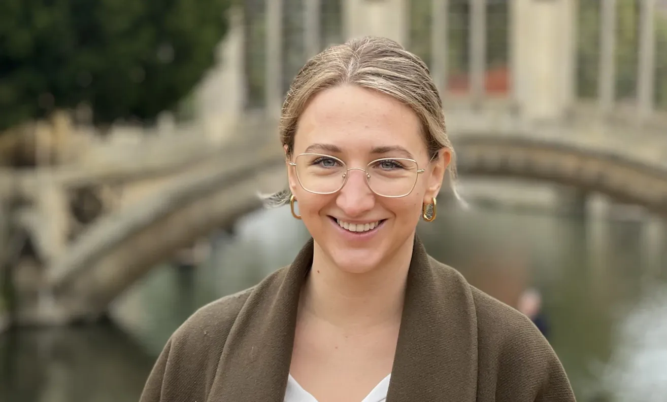 A woman with short blonde hair wearing glasses and a brown coat stands smiling near a bridge.