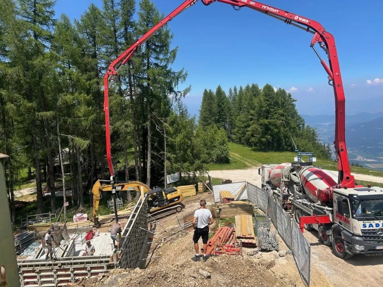 A construction site with a red crane and a concrete mixer truck. Workers are pouring concrete into a structure. The site is surrounded by trees.