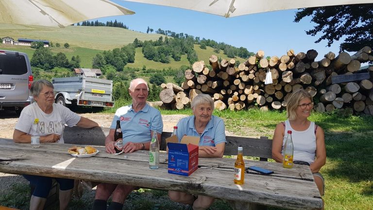 Zwei ältere Menschen sitzen auf einer Picknickbank mit Getränken und Essen. Ein großer Stapel Holzscheite steht hinter ihnen. Ein LKW ist in der Nähe geparkt.