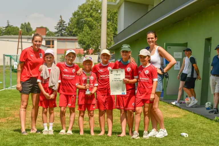 A group of young soccer players in red uniforms pose with a trophy and a certificate. They are standing on a green field with two adult women, one holding a watch. Trees and a building are in the background.