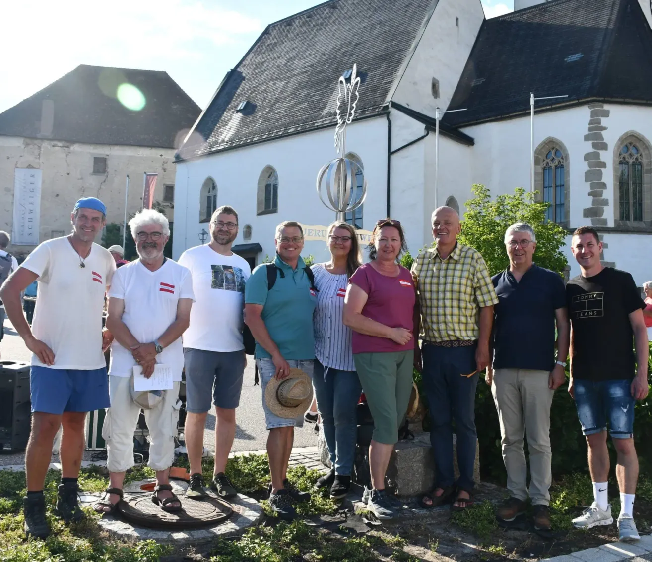 Eine Gruppe von Menschen steht vor einer Kirche und lächelt für ein Foto. Sie sind lässig in verschiedenen Farben gekleidet. Die Kirche hat ein schwarzes Dach und eine Statue hinter ihnen.
