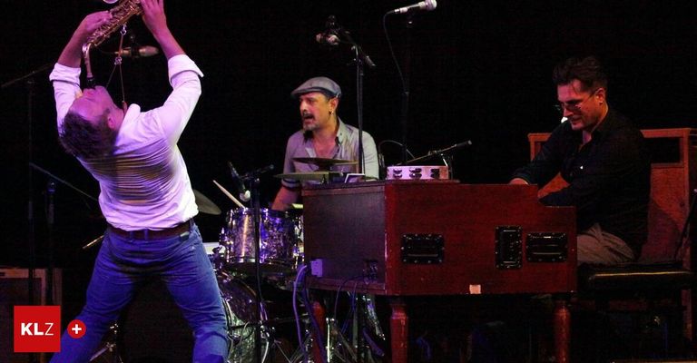 Eine Band auf der Bühne mit einer Person, die Saxophon spielt, einem Schlagzeuger mit einer Becken und einem Pianisten, der vor einem Klavier sitzt.