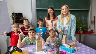 Eine Gruppe von Kindern und zwei Frauen lächeln in einem Klassenzimmer. Sie halten Papiertüten mit Lavendel und einem Schlüssel.