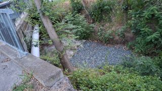 Eine kleine Kiesgrube, umgeben von dichter Vegetation und einer Betonstruktur. Die Grube ist mit Kies gefüllt und scheint ein Außenbereich für Drainage zu sein.