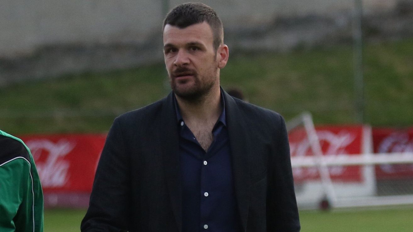 A man with a beard and mustache wearing a dark suit and blue shirt is standing on a grassy field. He looks serious.