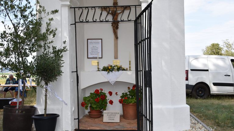 Ein weißer Kapelleneingang mit einem Metalltor, einem Kreuz darüber und einem Schild für Dorfmeister Kapelle erbaut 1806. Vor dem Eingang stehen Blumentöpfe und Blumen.