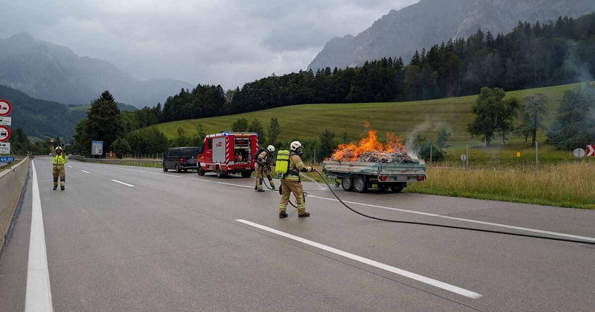 Firefighters extinguish a burning trailer on the side of a mountain road with a black van and a fire truck nearby.