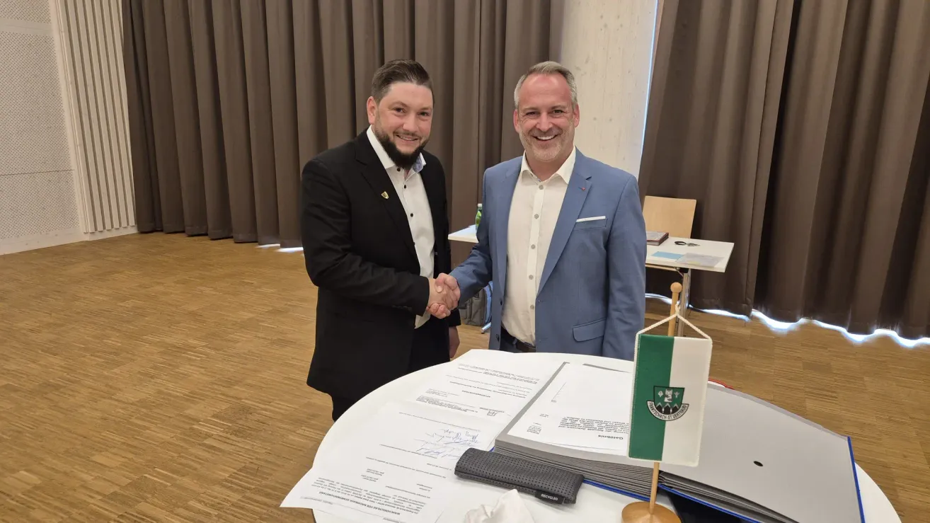 Two men in formal attire, shaking hands, standing before a desk. They are smiling and appear to be celebrating a formal agreement. Papers, a small flag, and a pen are on the table.