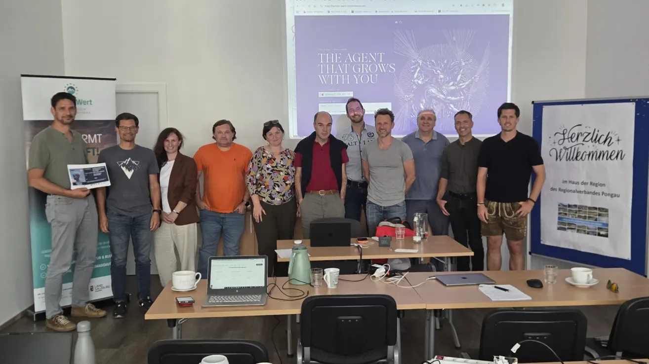 A group of people stands in a meeting room with a projector screen. The screen displays a website with 'The agent that grows with you.' The people wear casual attire, and laptops, glasses, and cups are on the desk.