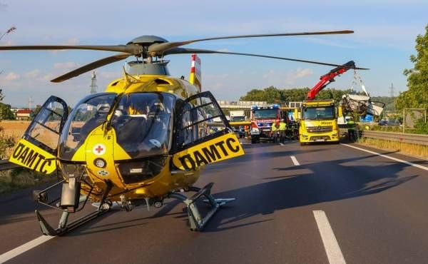 A yellow helicopter is on the side of the road. DAMTC is written on it. An ambulance is parked nearby, and a crane is lifting an object.