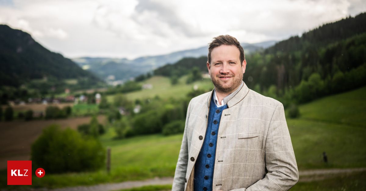 A man in a suit stands smiling in a field with a view of mountains.