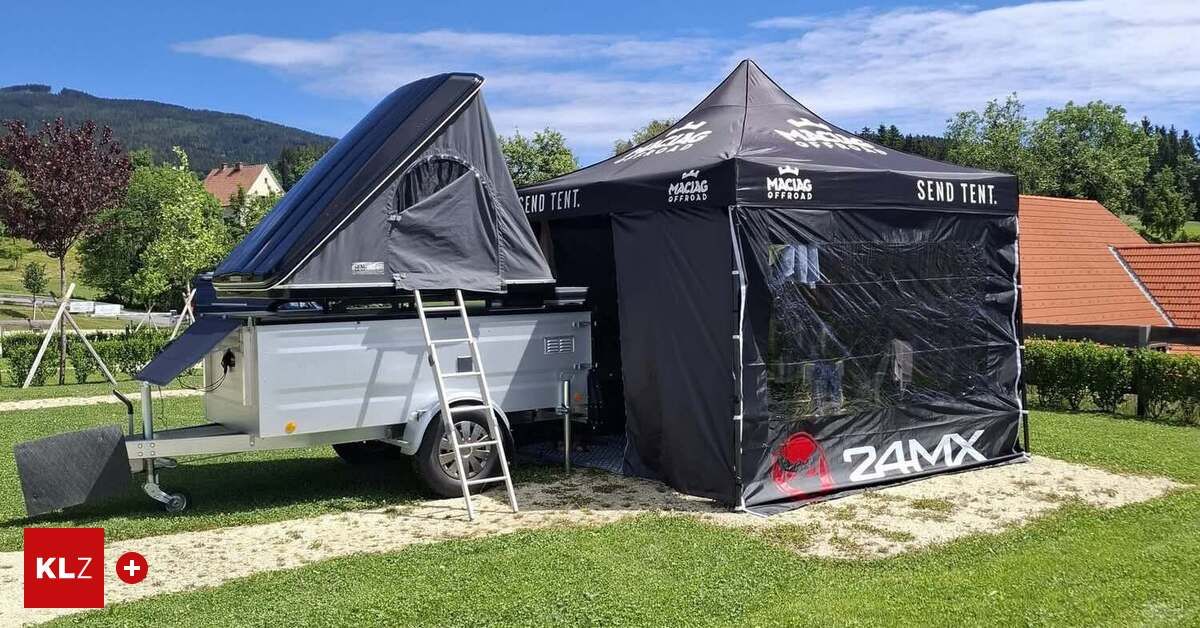 A black tent with a ladder attached stands next to a silver trailer with an open top, both on a grassy field under a blue sky.