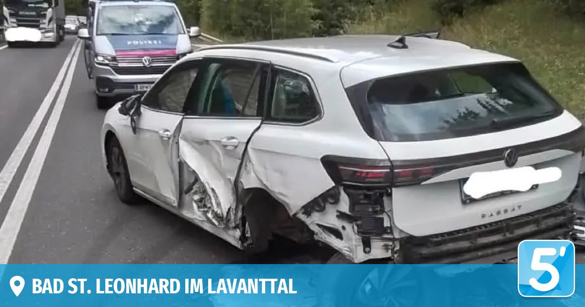 A white car with severe rear-end damage is parked on a road, with a police car behind it. The car's back bumper is crushed, and the trunk lid is open.
