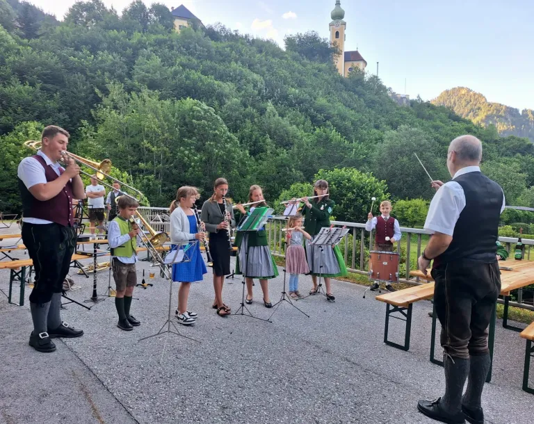 Eine kleine Band von Kindern in traditionellen Kleidungsstücken spielt Instrumente in einem Bergdorf. Der Dirigent trägt eine Weste und Stiefel.