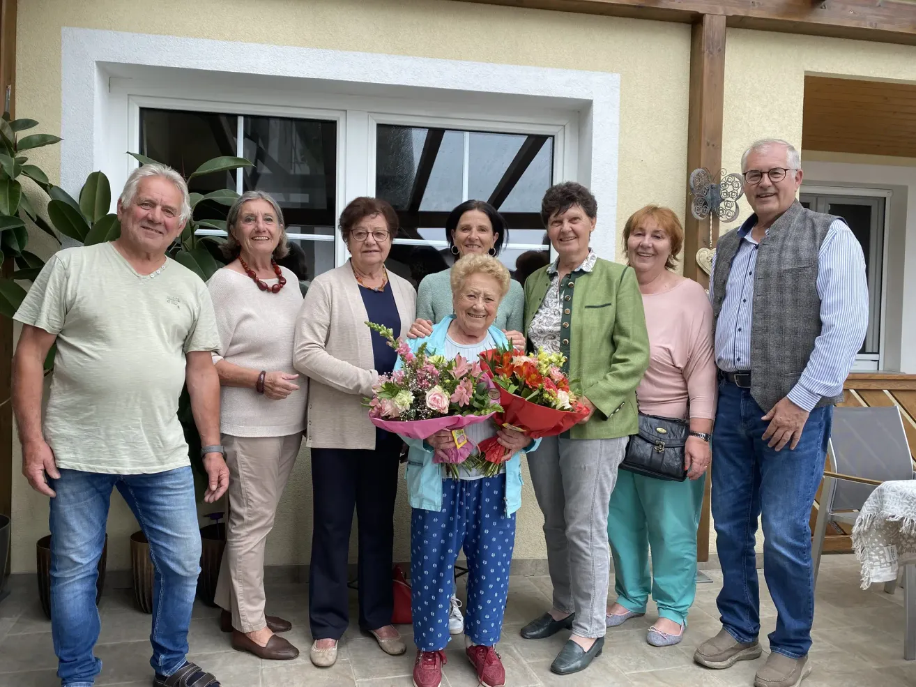 A group of people are standing outside near a window. One woman holds flowers. The others smile and look at the camera.
