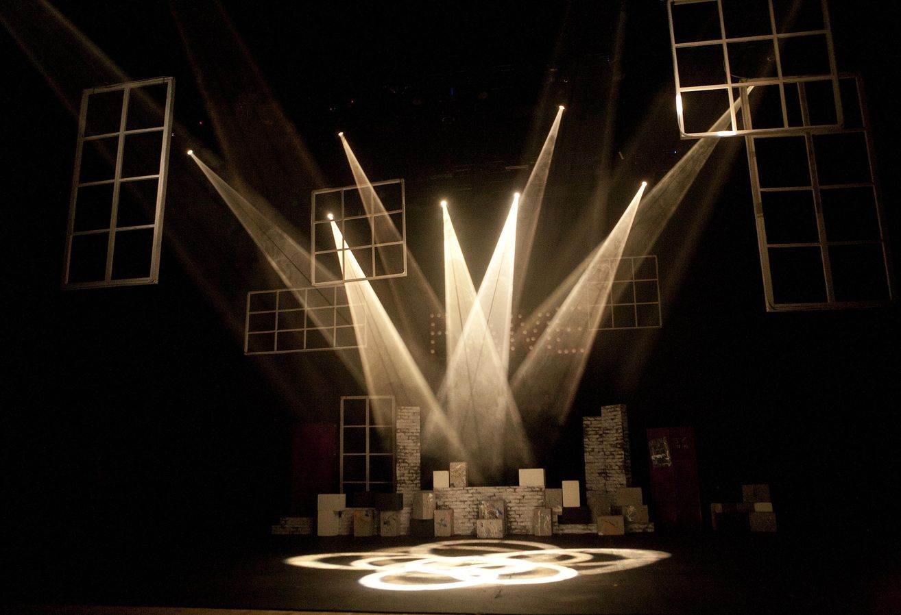 A stage set with spotlights illuminating a cityscape, featuring various structures and buildings with a circular design on the stage floor.