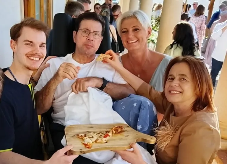 A family sits together outdoors, smiling. A man in glasses eats pizza from a wooden board. A woman in a blue dress smiles at him. Another woman holds the board.