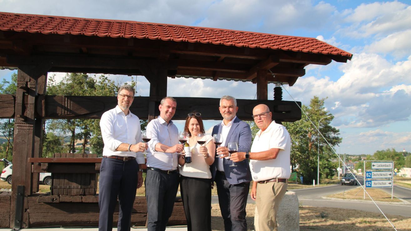 Five individuals are standing under a gazebo, smiling and holding wine glasses. One person is holding a bottle. They appear to be posing for a photo.