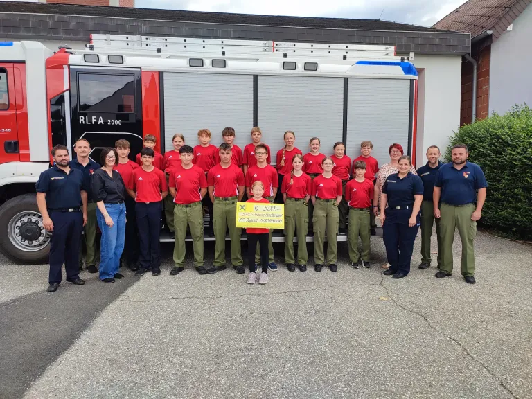 Eine Gruppe von Menschen, darunter Kinder, steht vor einem Feuerwehrauto. Ein Kind hält ein Schild mit 500 Euro.