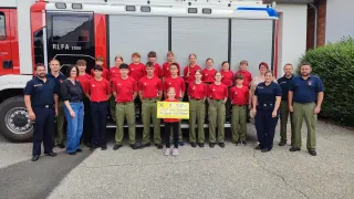Eine Gruppe von Menschen, darunter Kinder, steht vor einem Feuerwehrauto. Ein Kind hält ein Schild mit 500 Euro.