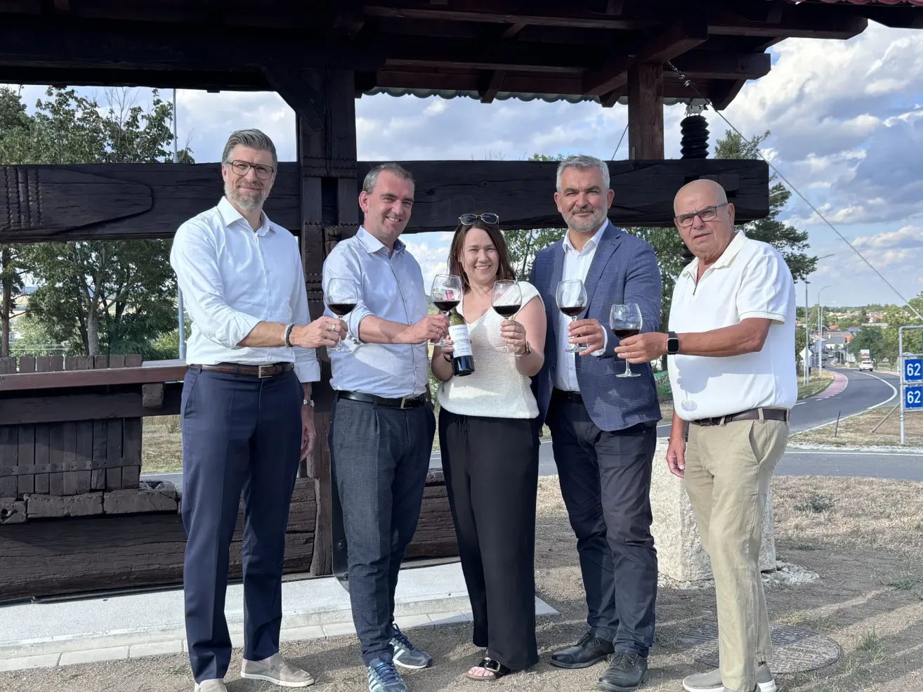 Five people, three men and two women, stand in front of a wooden structure, all holding wine glasses. They are dressed formally, suggesting a celebratory or professional event.