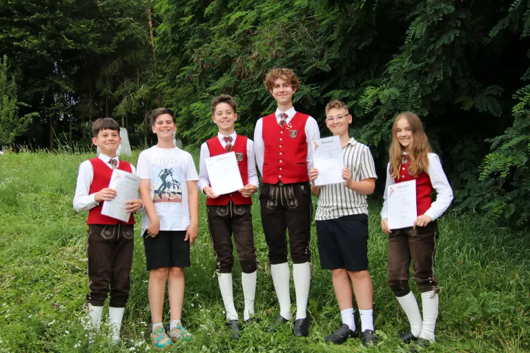 A group of young people are standing in a field holding certificates. They are dressed in traditional clothing and appear to be posing for a photo.