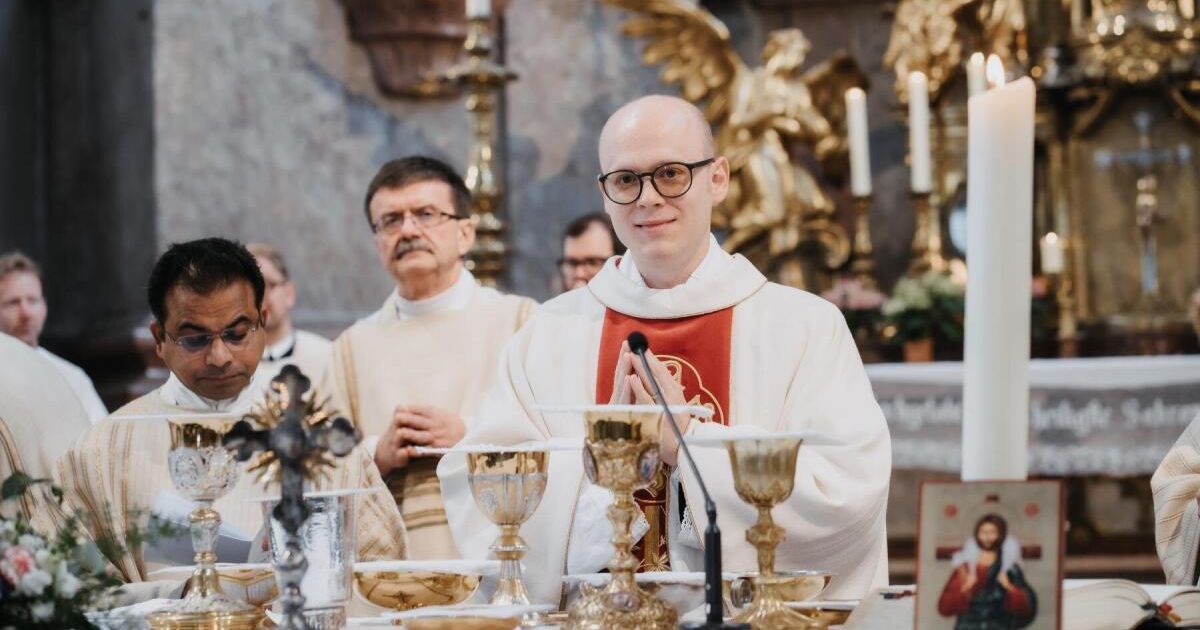 A priest stands in front of an altar with chalices and candles, wearing glasses and a white and red vestment. Behind him, two other men in clerical attire look on.