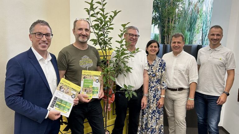 Five people stand near a tree, smiling. They hold pamphlets with green text. The man on the left wears glasses. A yellow container is behind them.