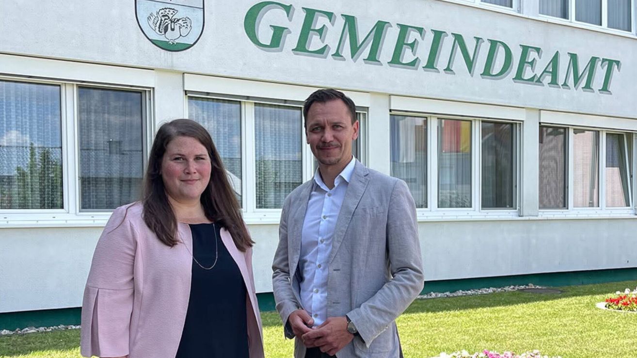 A man and a woman stand in front of a building with a sign reading Gemeinde. The woman wears a pink jacket and the man a gray blazer. Both smile and appear to be posing for a photo.