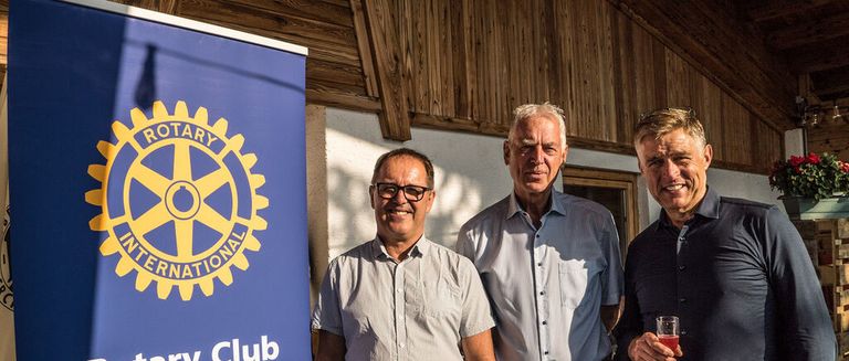Three men stand close together in front of a wooden wall. A man in the middle smiles, while the man on the right holds a glass. The blue banner behind them features the Rotary International logo.