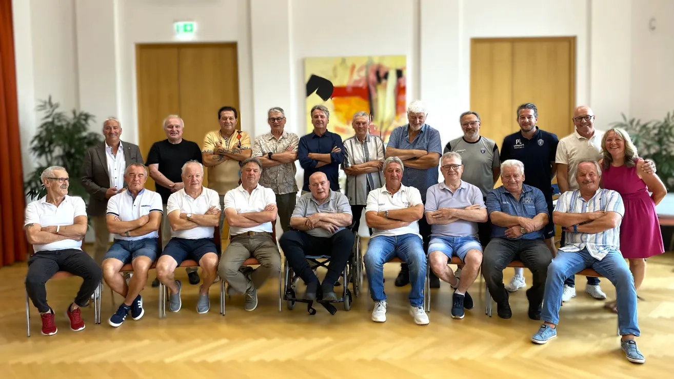 Eine Gruppe von Männern, einige sitzend und einige stehend, posiert für ein Foto in einem Raum mit Holzböden und einem großen Gemälde.