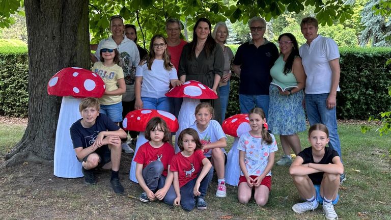 A group of people, including children and adults, stand and crouch under a tree. Some wear mushroom hats. One woman holds a book. They seem to be posing for a photo.